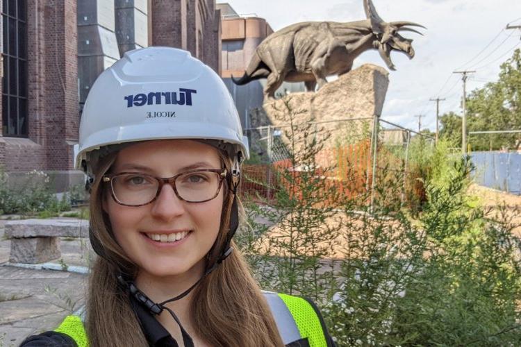 Nicole Zalewski, wearing glasses and a construction helmet, stands in front of an old brick building with a statue of a triceratops out front.